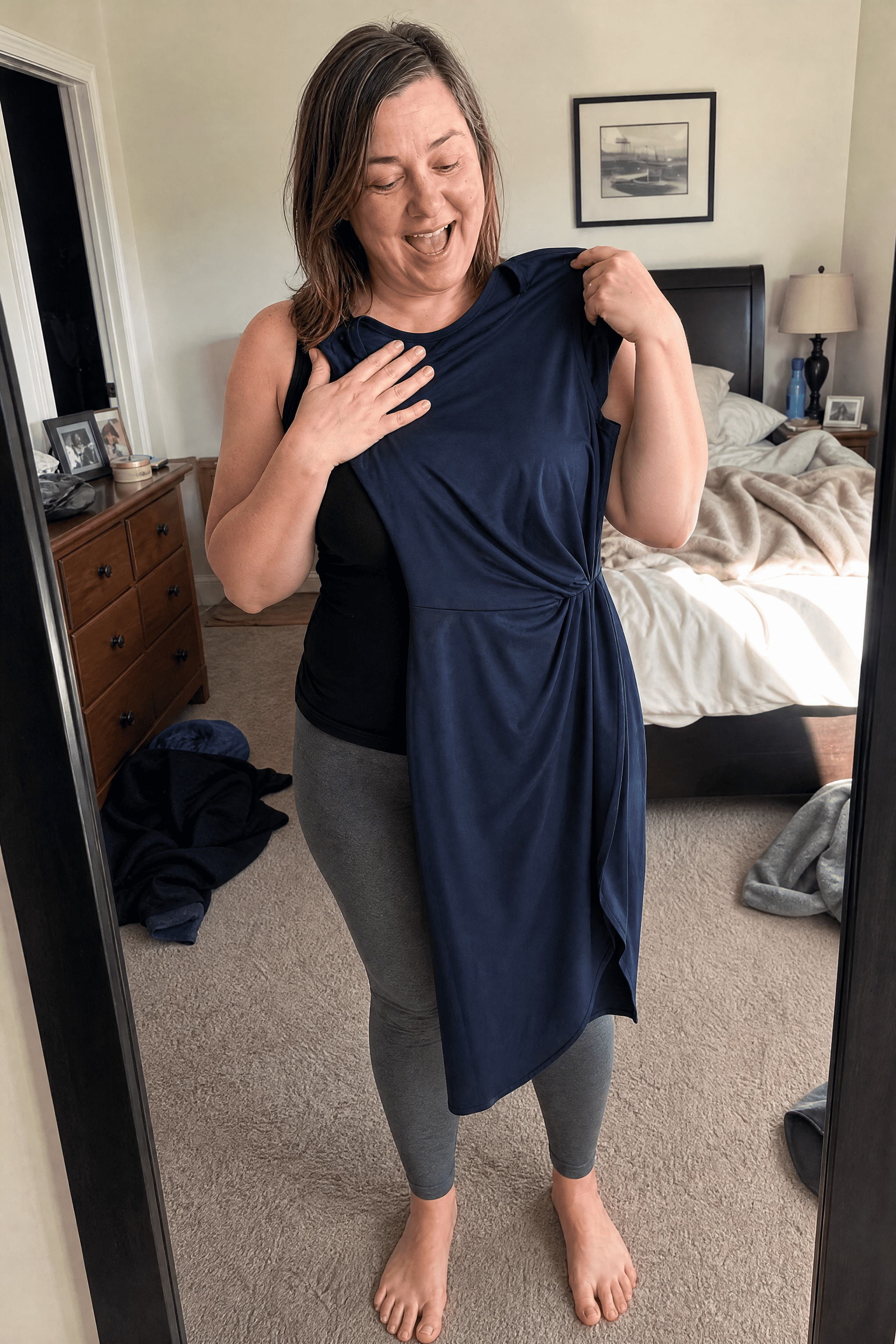 Tania holding a fitted dark dress against herself in front of a mirror, smiling in happy disbelief